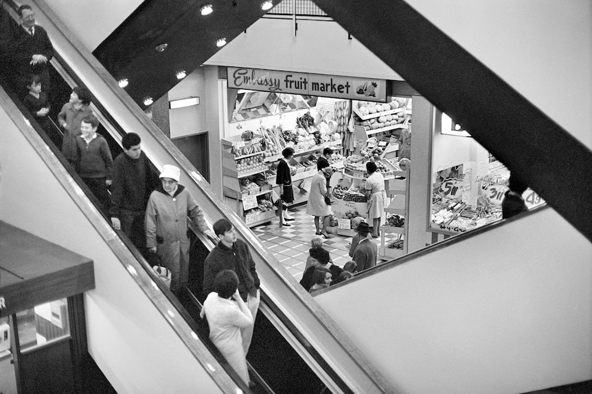 A back and white image of people on an escalator in a busy shopping centre.