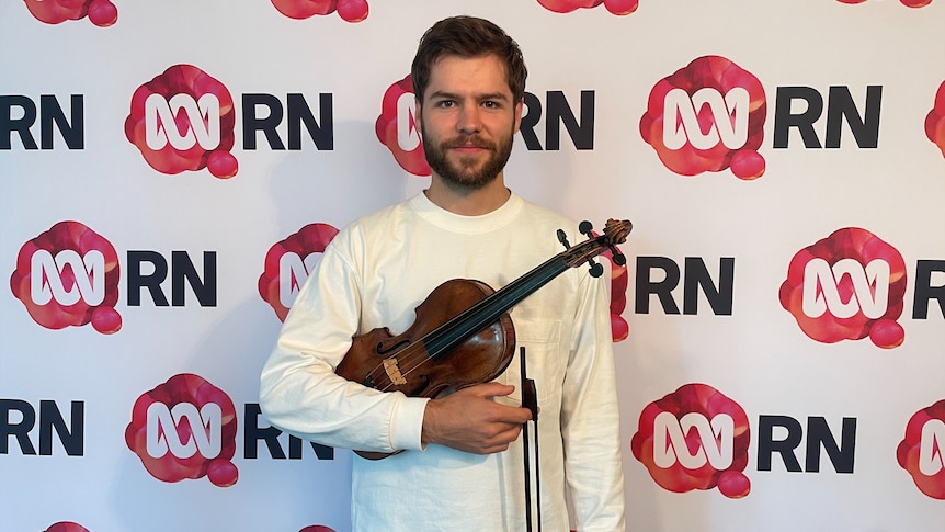 man holds a violin and gives a slight smile to the camera, he stands in front of a wall with RN branding 