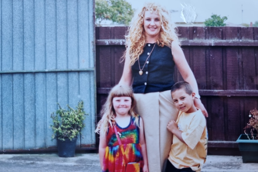 An old coloured photo of a blonde woman with perm holding two smiling children by her side.