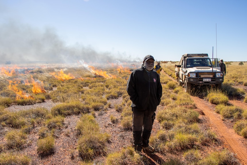 A man stands in desert landscape as small fires burn behind him