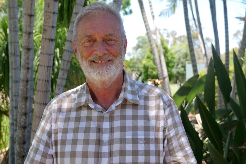 Dr Alistair Melzer looking at the camera smiling, white and brown check shirt, trees and garden behind.