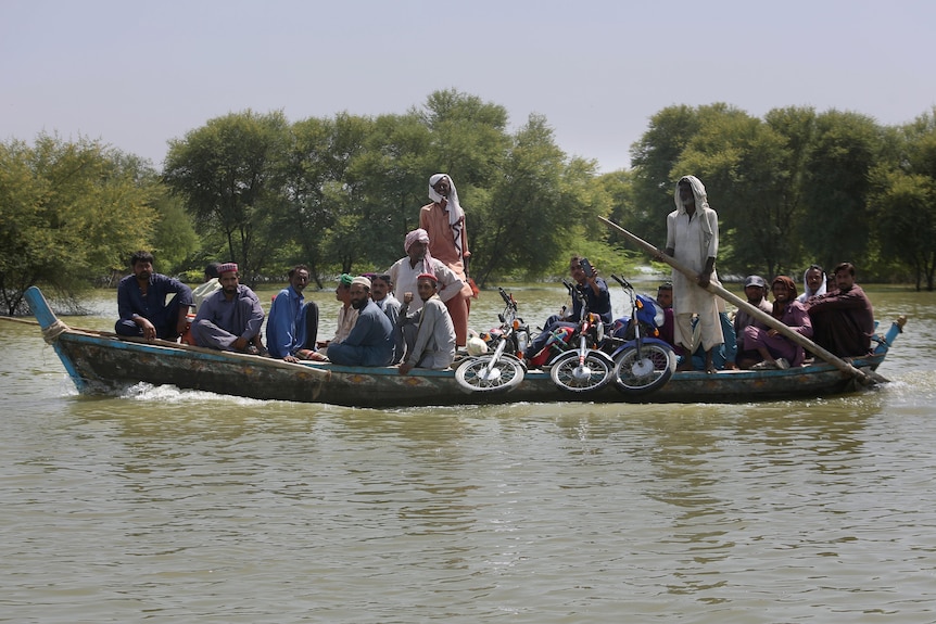 About 19 men and several motorcycles are tightly packed on a low boat making its way past some trees.