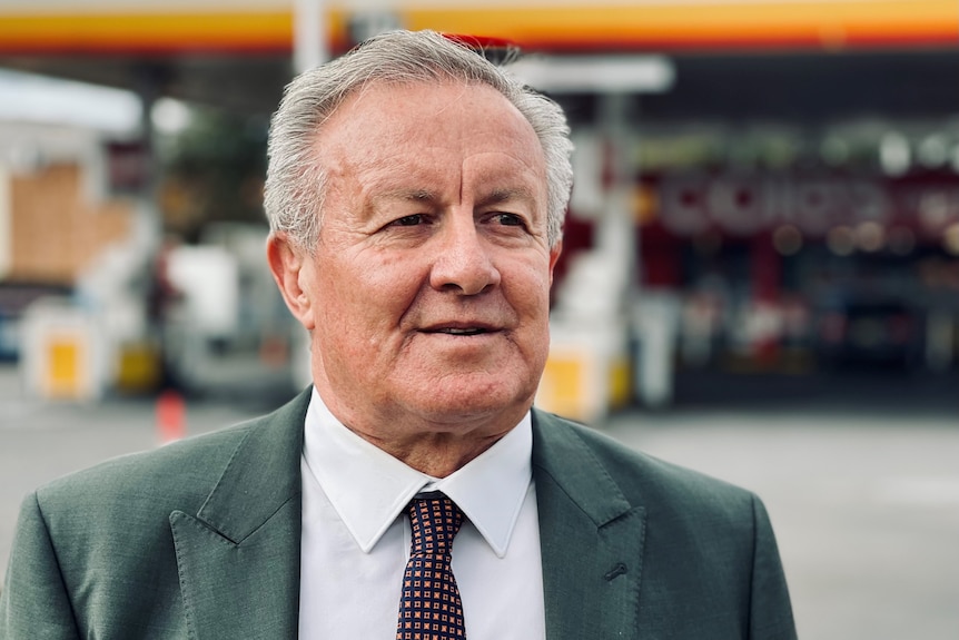 An older white man wearing a suit stands in front of a Shell petrol station.