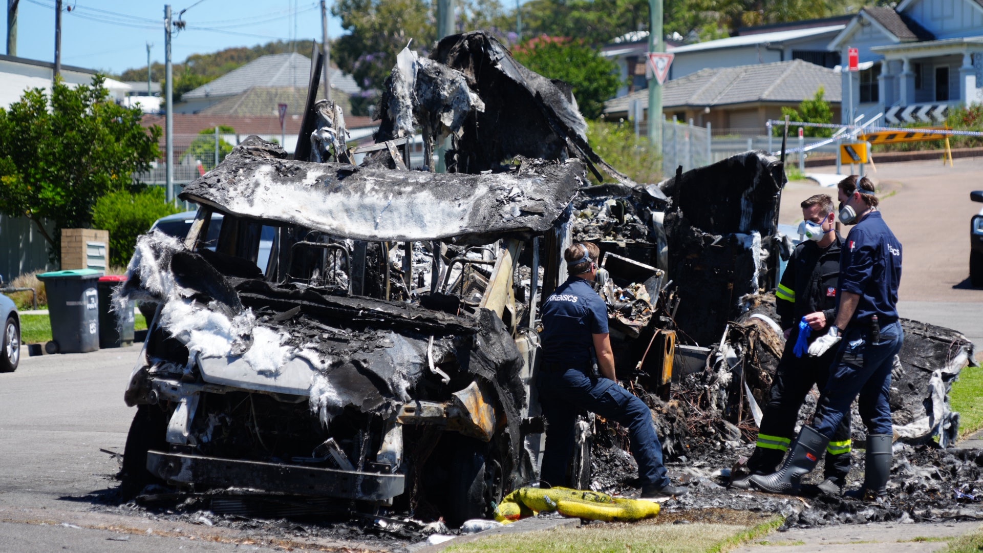 A severely burnt motor home which has been destroyed on a street with rescue crew members around it.