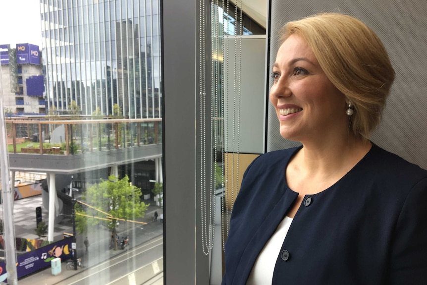 A smiling woman with short blonde hair stands at a window in an office building.