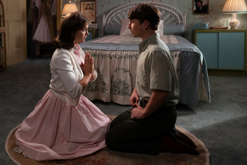 A white girl in a pink dress with dark hair kneels in prayer in front of white boy with grey shirt and dark hair in a bedroom.
