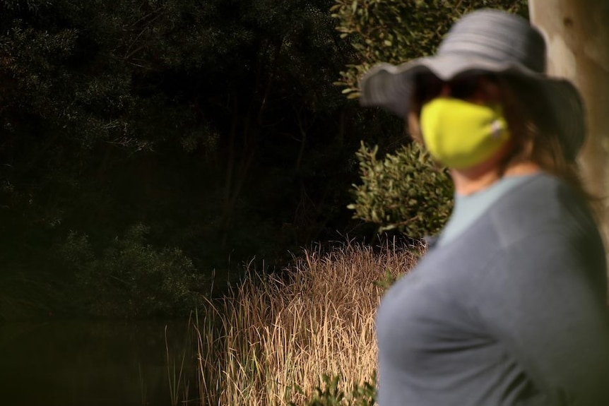 A woman wears a hat and a bright yellow face mask. She's standing by a creek in the bush.