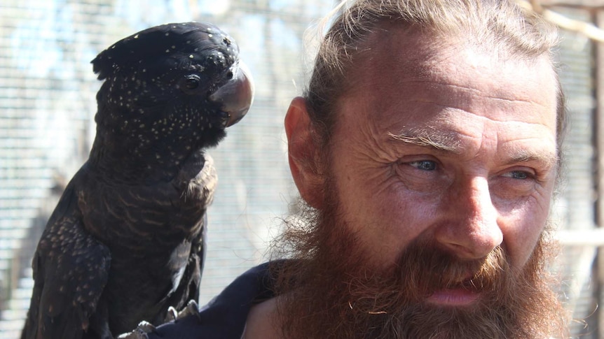 Damian Stanioch has a beard and stands with a black cockatoo on his shoulder