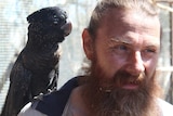Damian Stanioch has a beard and stands with a black cockatoo on his shoulder
