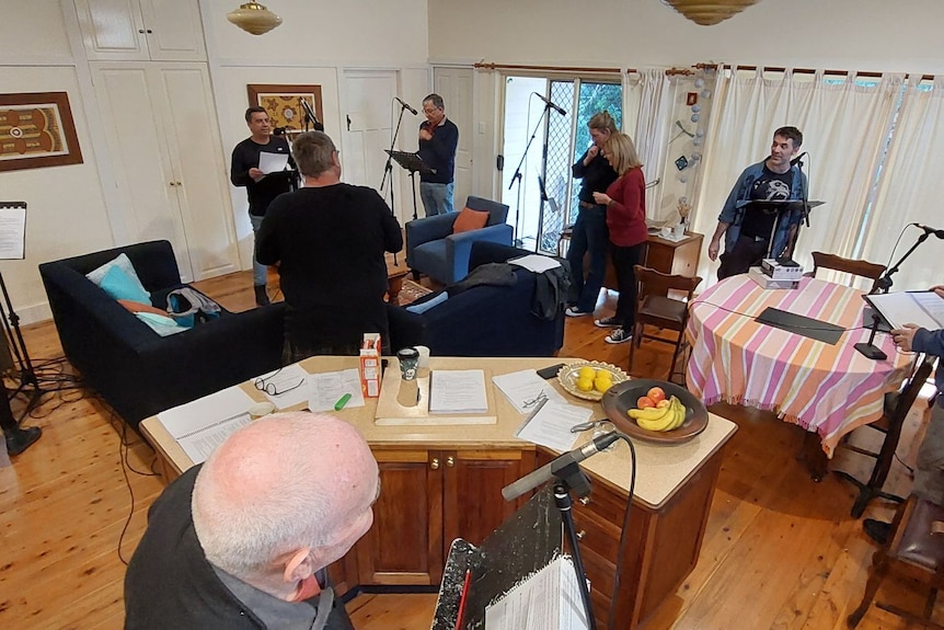 A group of people stand at music stands and microphones, spaced around a living area.