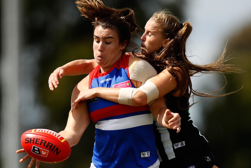 An AFLW player holds the ball free as she is tackled from behind by a defender.