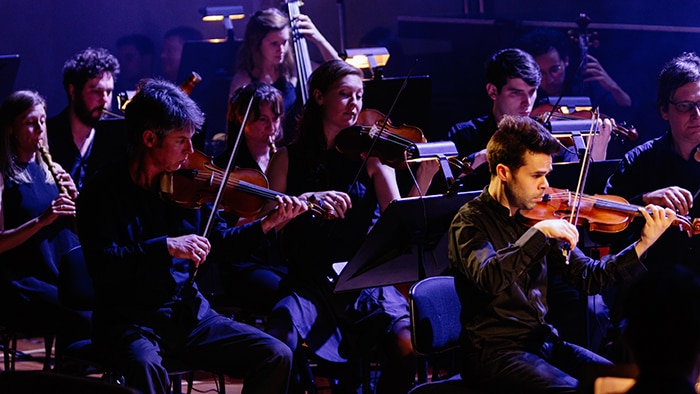 The string section of Pinchgut Opera's Orchestra of the Antipodes performing on stage with moody blue lighting