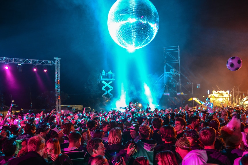 A crowd of thousands gather on an outdoor dancefloor under a giant disco ball as lights beam into the sky