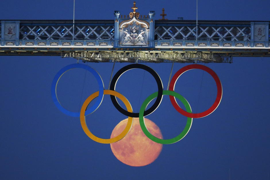 The full moon rises through the Olympic Rings hanging beneath Tower Bridge in London.