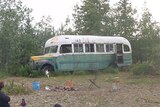 An old school bus sits among trees. Campers sit nearby.