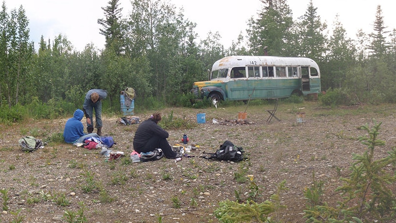 An old pale green school bus sits among trees. Campers sit nearby.