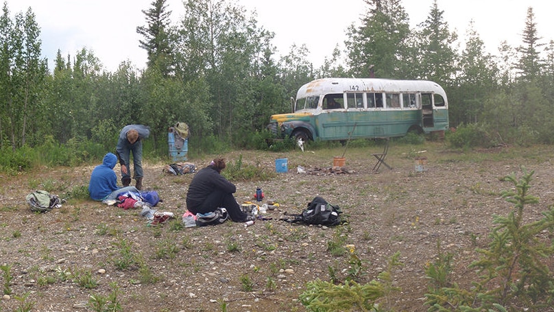 An old school bus sits among trees. Campers sit nearby.