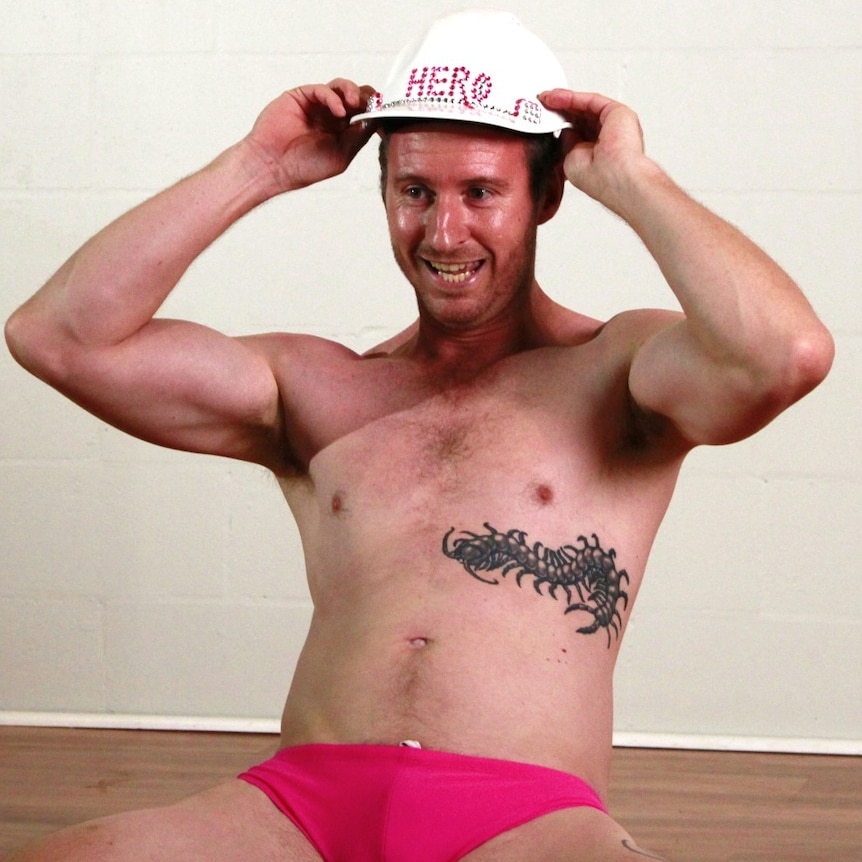 Man holds white hard hat with hero written on it, while kneeling on the floor wearing pink speedos