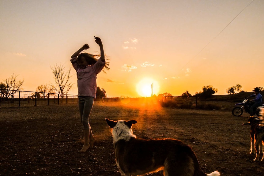A young girl dances in the dirt at sunset with her dog nearby