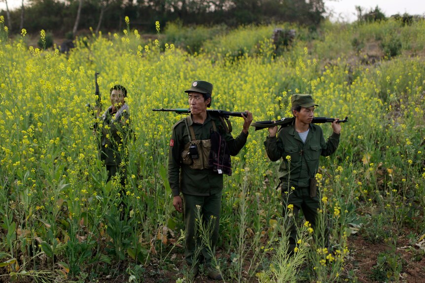 Three MNDAA soldiers with guns stand amongst tall wildflowers. 