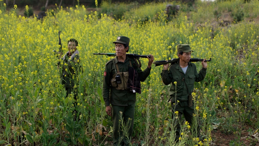 Three MNDAA soldiers with guns stand amongst tall wildflowers. 