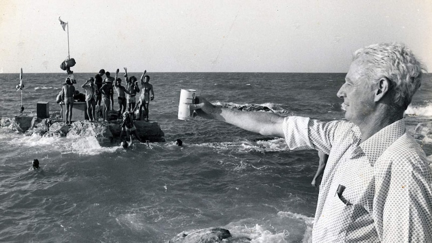 An archival photo of a man raising a can of beer to a group of people on a rock.
