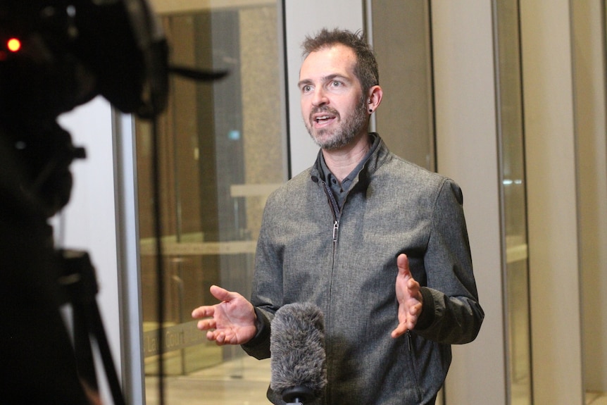 Middle-aged white man in a grey jacket speaking in front of TV camera and microphone