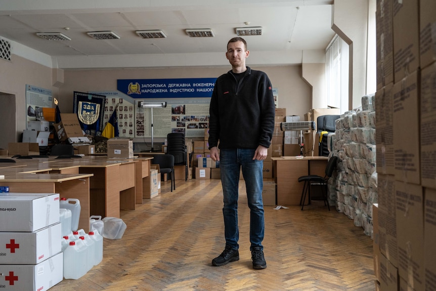 A skinny man in jeans and a jumper stands among cardboard aid boxes branded with red crosses