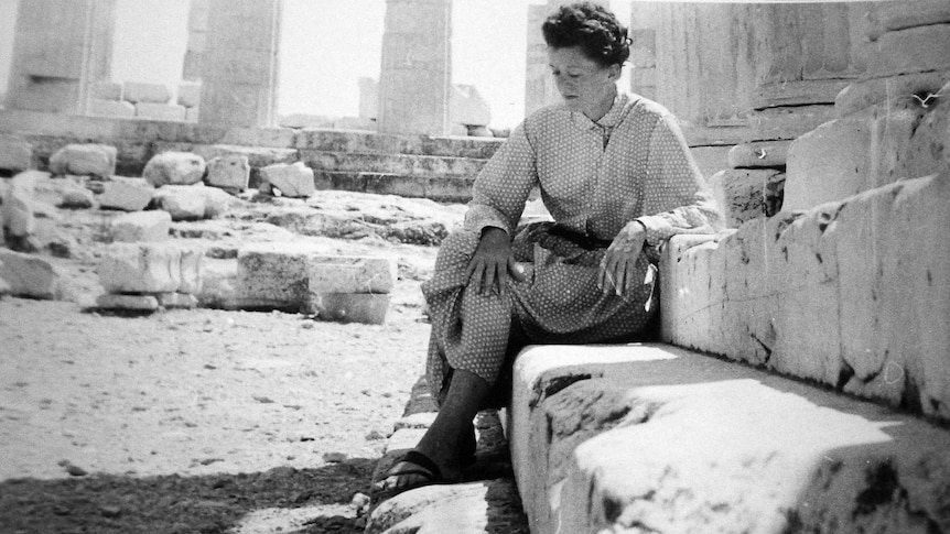 A woman wearing a long-sleeved spotted dress and sandals sits in the ruins of an ancient Greek building.