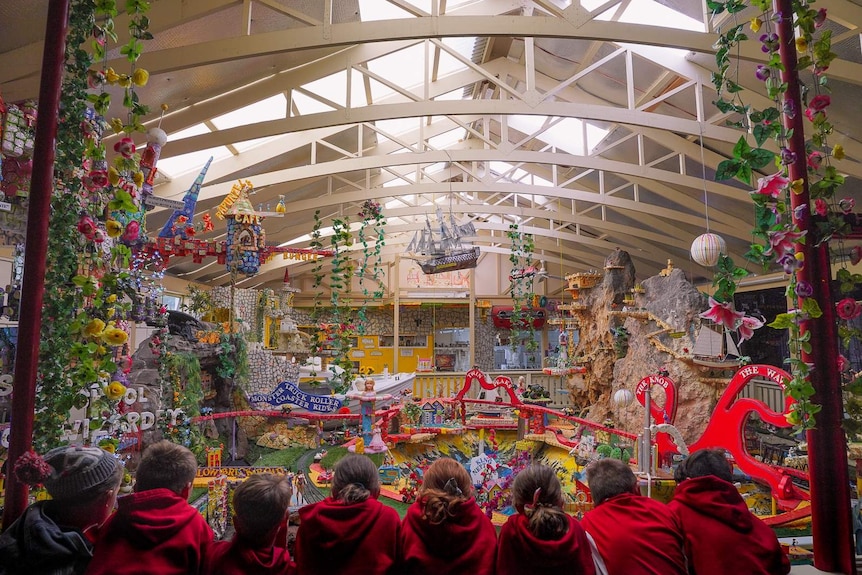A row of children in red school jumpers look out over a large indoor theme park filed with colourful model trains and displays.