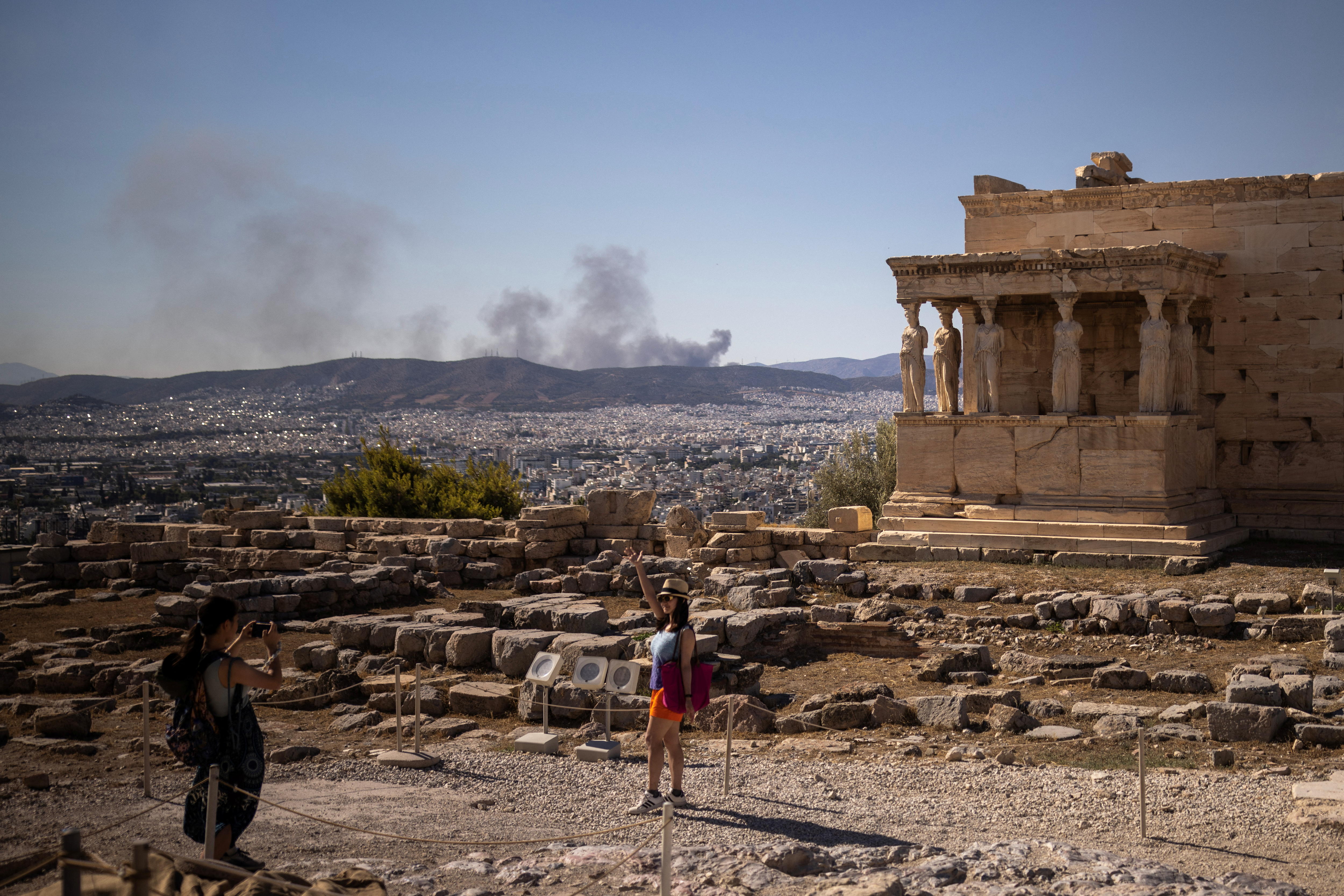 A tourist poses in front of a Greek monument while wildfires burn in the background