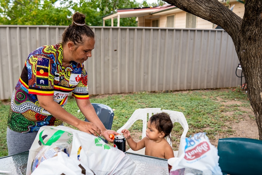Indigenous woman in a shirt with colourful traditional art feeds a young boy in a white plastic chair a strawberry.