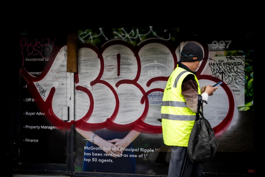 A person wearing a high visibility vest walks in front of a real estate sign that has been painted over with graffiti.