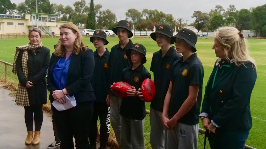Three women, and five school students in green uniforms holding AFL footballs, stand in a group at a football oval.