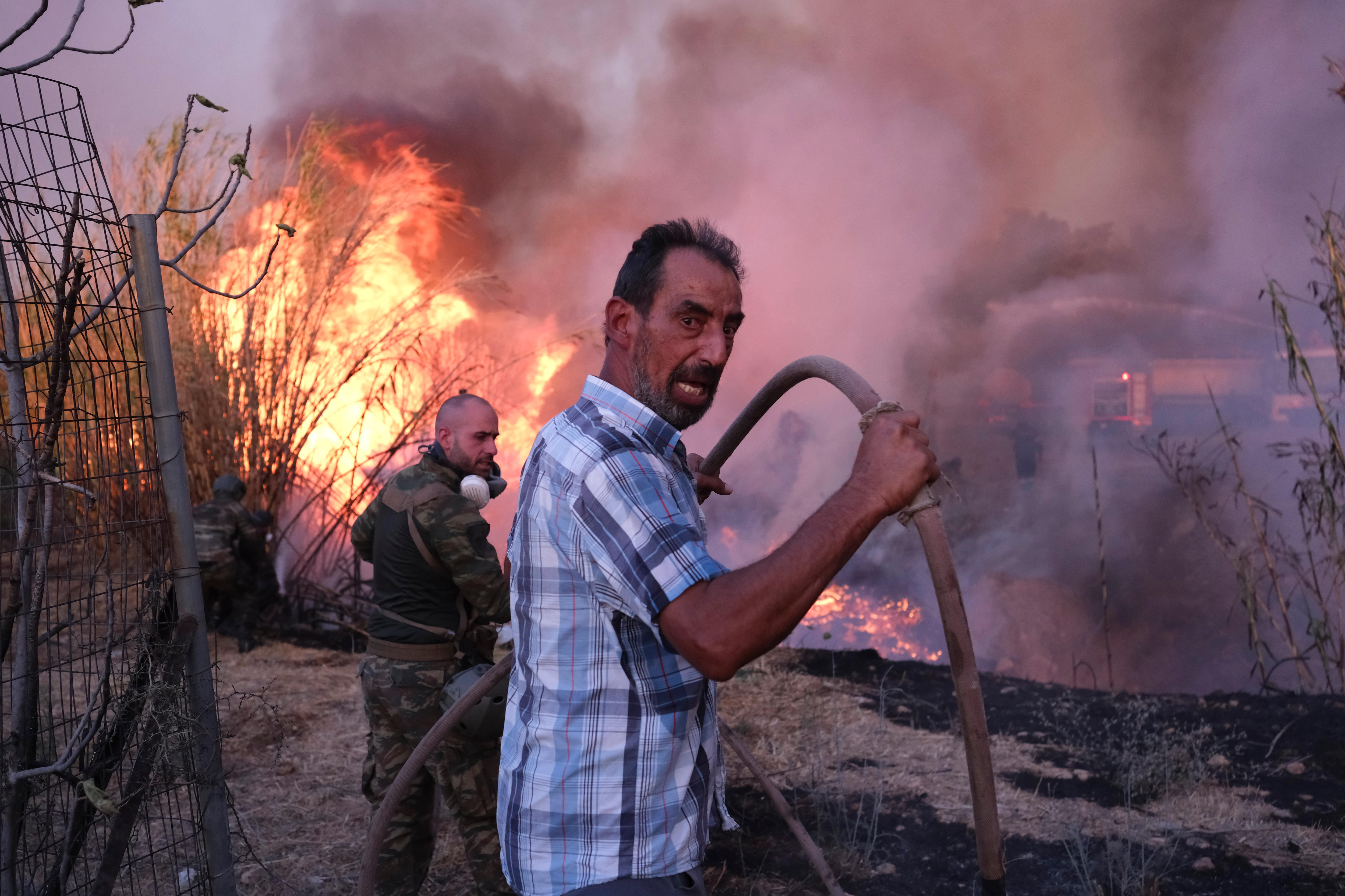 Two men hold a firehose in front of a fire on shrubland.