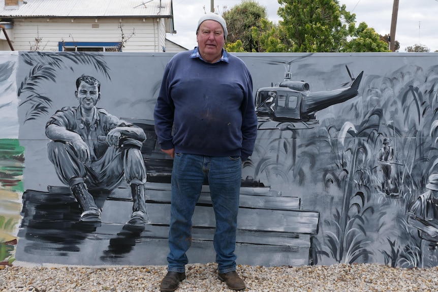 Un homme en bonnet se tient devant une peinture murale grise représentant un jeune homme assis sur un véhicule.