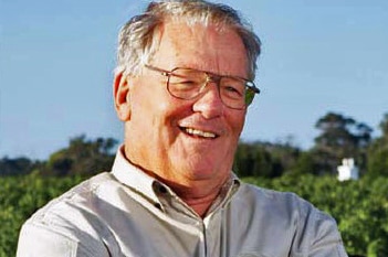 A head and shoulders shot of Michael Wright smiling outdoors in a vineyard wearing spectacles and a grey shirt under a blue sky.