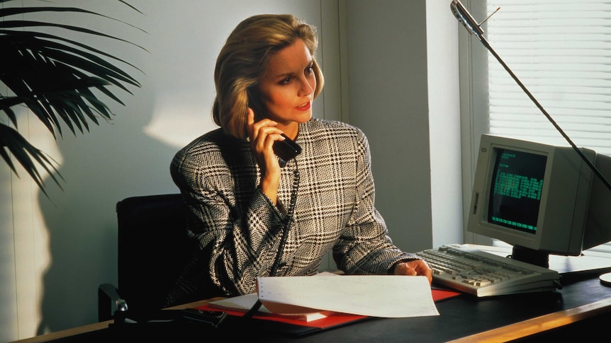 A woman in 1980s business attire sits at a desk with a telephone receiver to her ear.