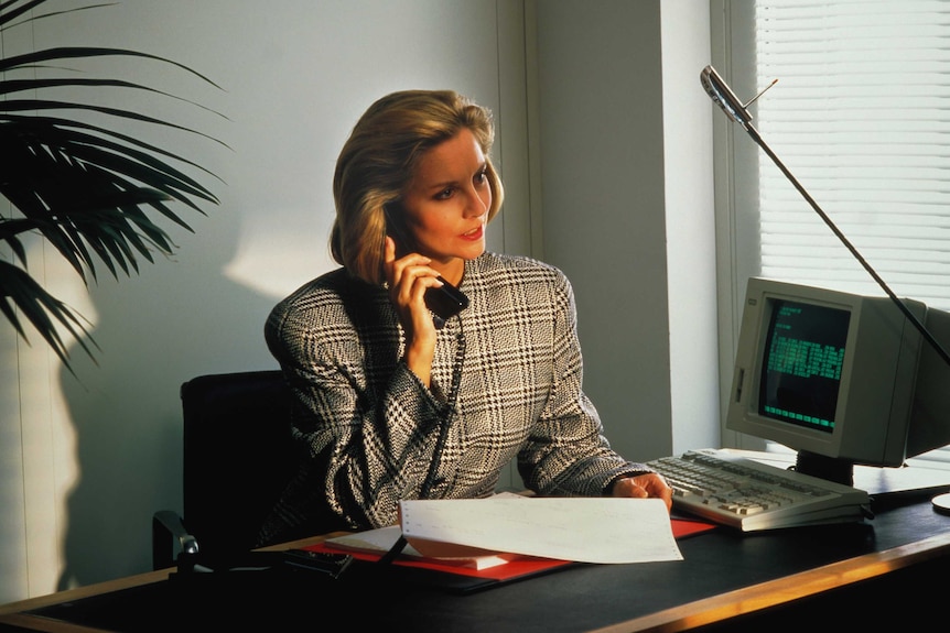 A woman in 1980s business attire sits at a desk with a telephone receiver to her ear.