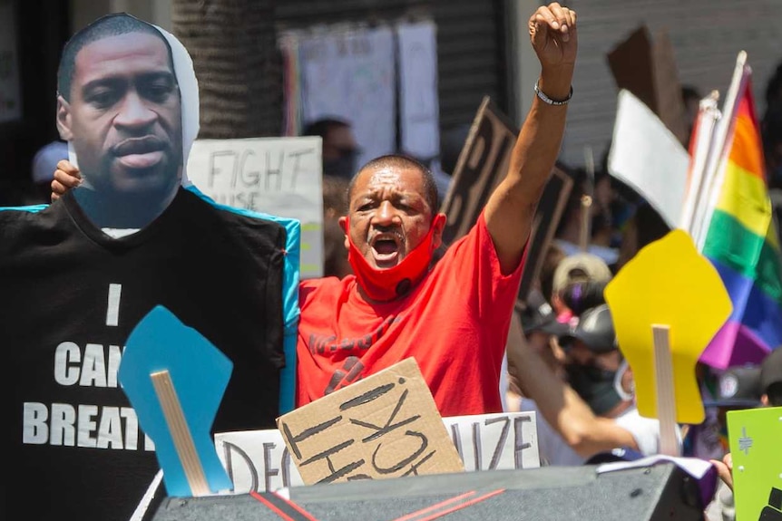 Man in red shirt with right hand raised stands amid a crowd protesting racial injustice.