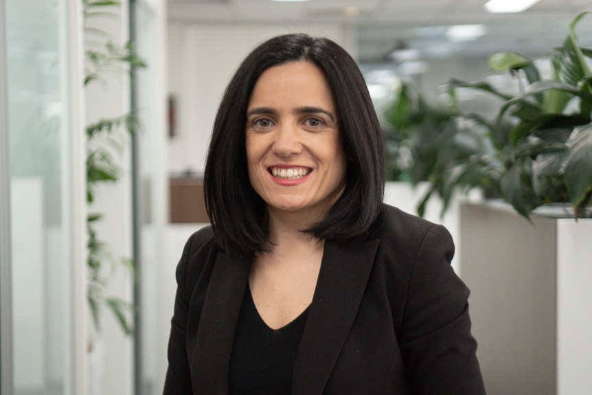 A woman wearing a black jacket smiles at the camera while standing in an office space.