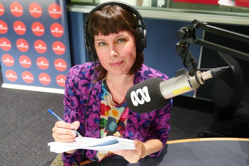 Cook wearing headphones behind microphone in studio holding pen and notepad.