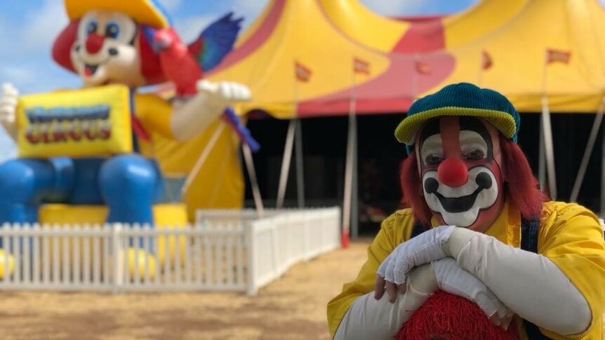 An adult and a child dressed as clowns stand outside a big top