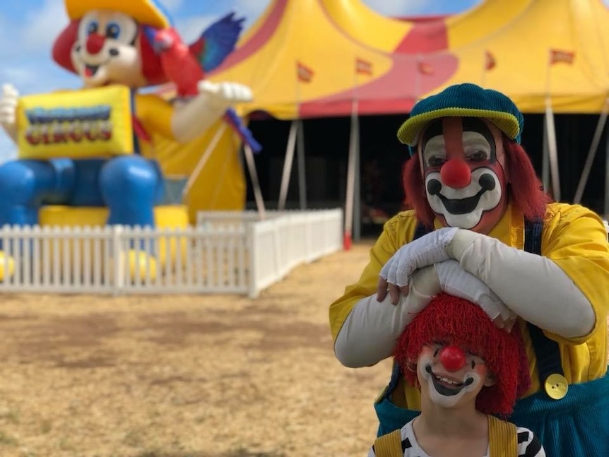 An adult and a child dressed as clowns stand outside a big top