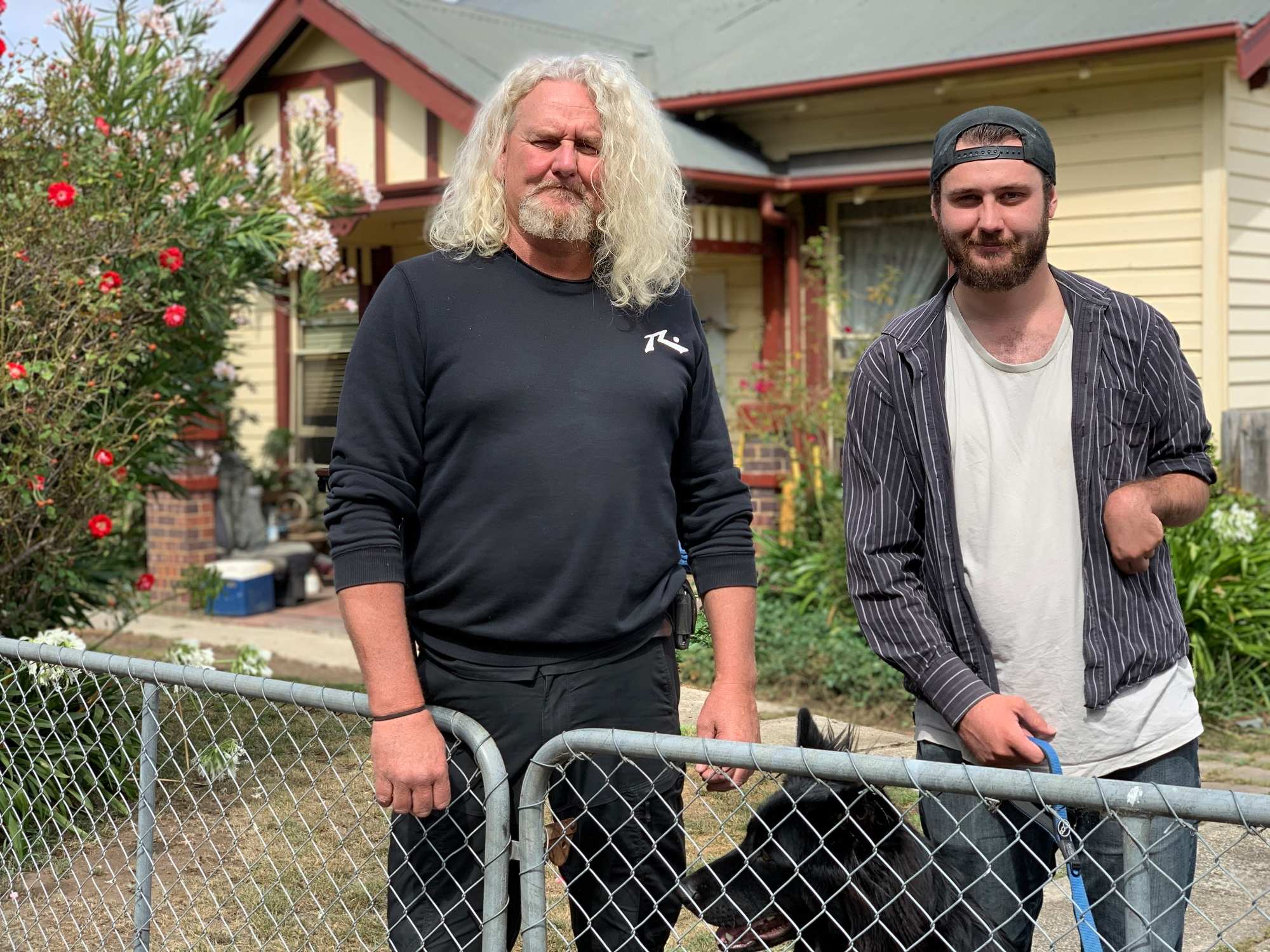 Two men stand behind a wire gate in front of a house.