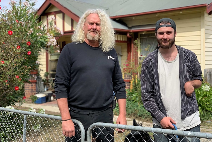 Two men stand behind a wire gate in front of a house.