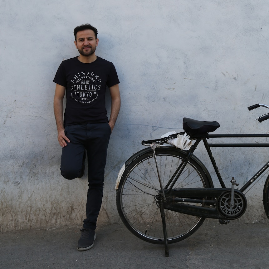 A young Hazara man in a black Tshirt standing in a street next to a bike.