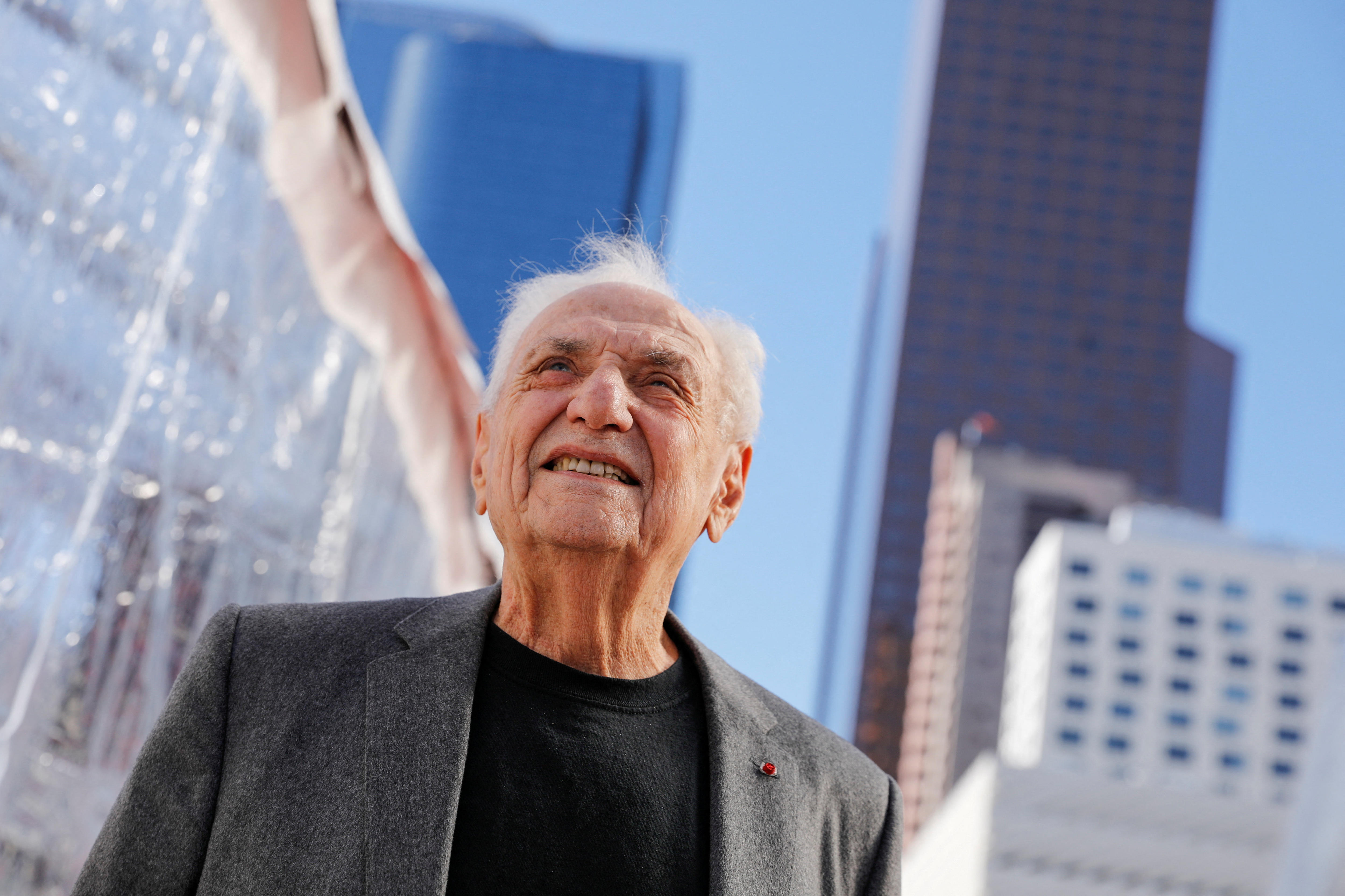 An older man with white hair smiles in the sun in front of a large, rippled steel building.