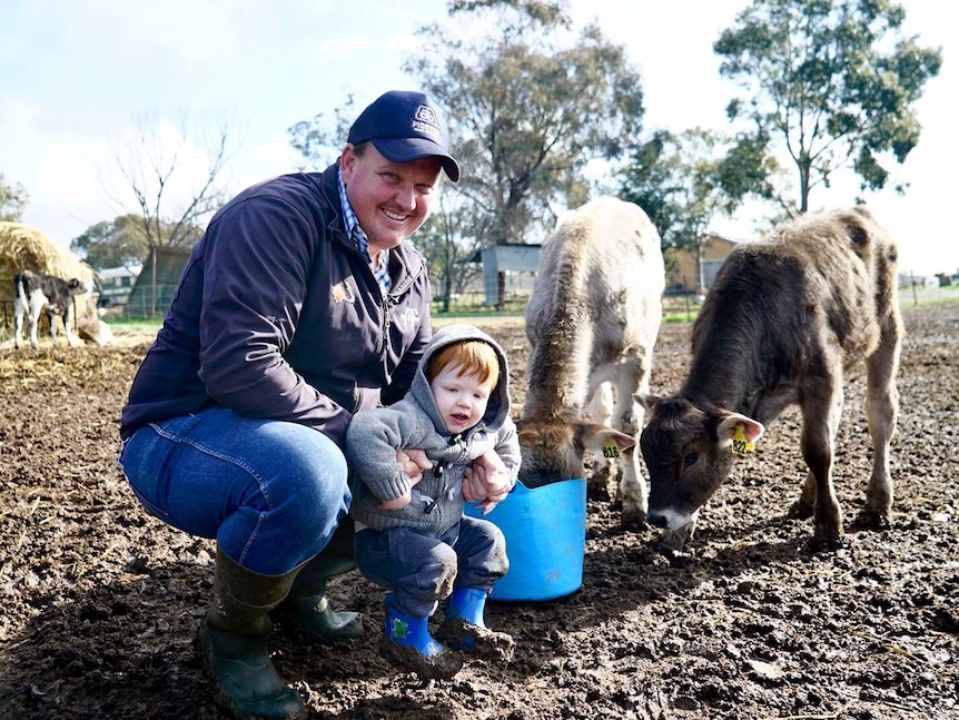 A smiling farmer kneels down in the mud, holding onto a little boy as a few young animals go about their business behind them.