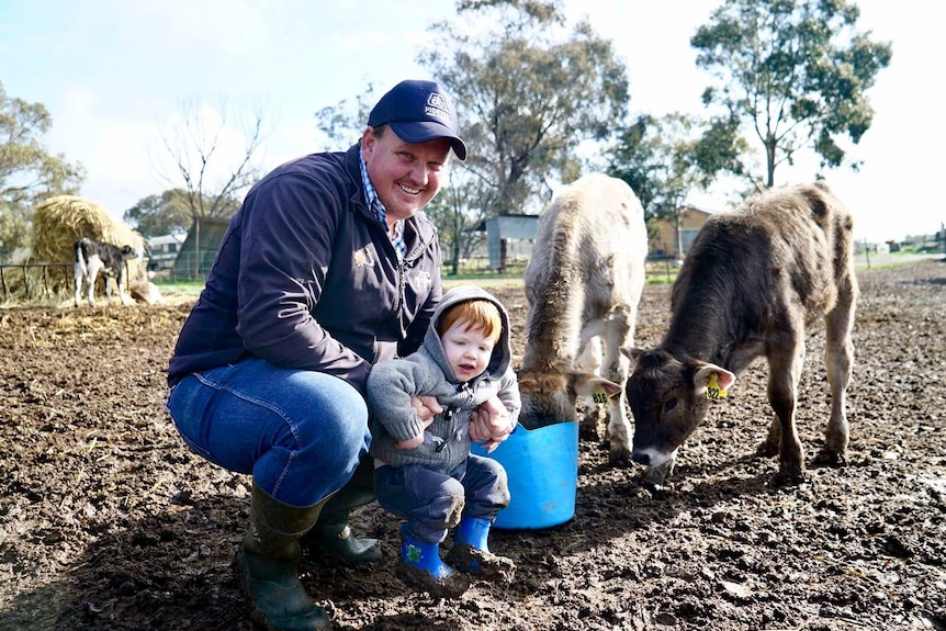 A smiling farmer kneels down in the mud, holding onto a little boy as a few young animals go about their business behind them.